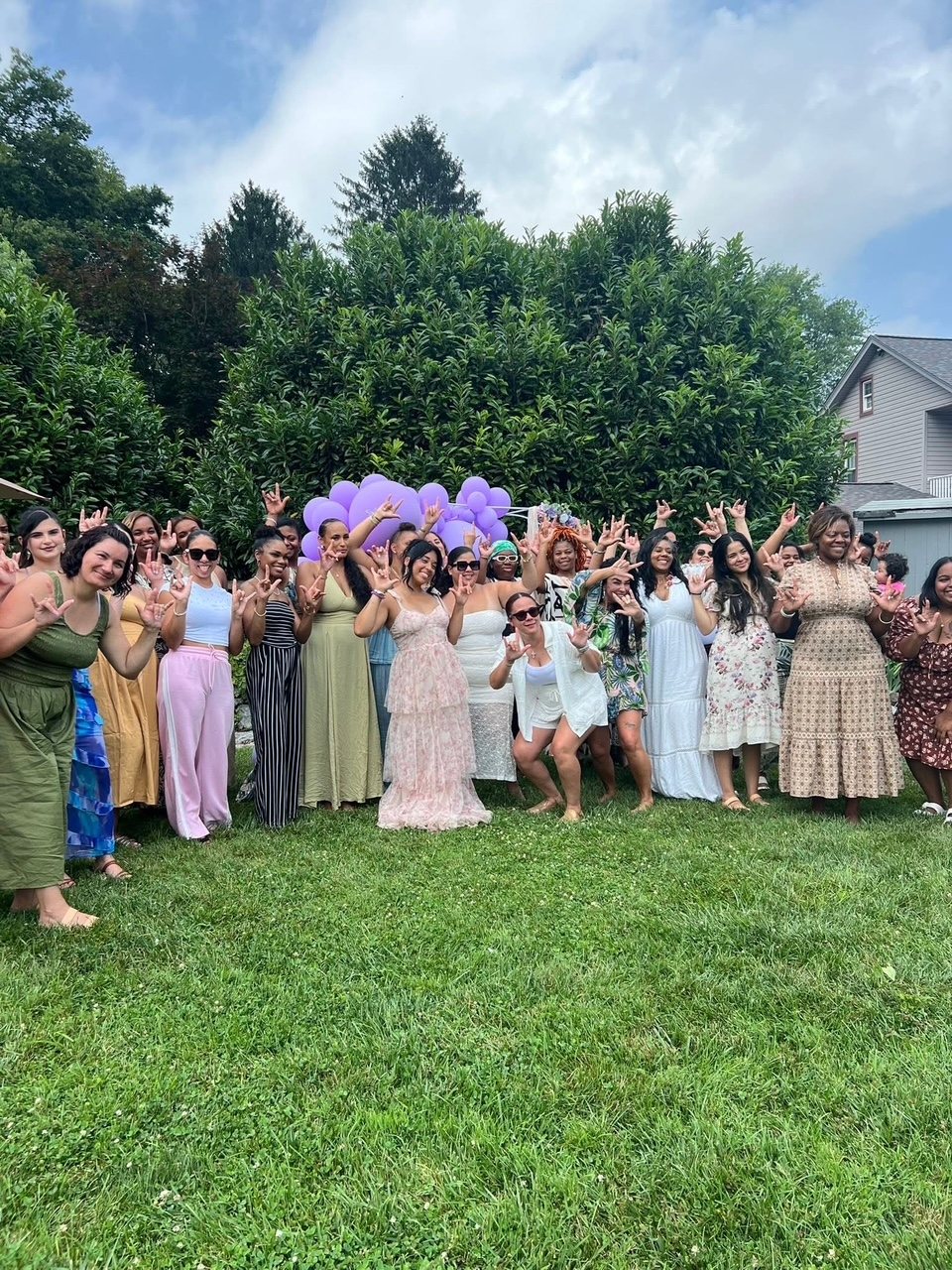 The women of MOMDAY on a lawn under purple balloons, every one of them signing I love you.